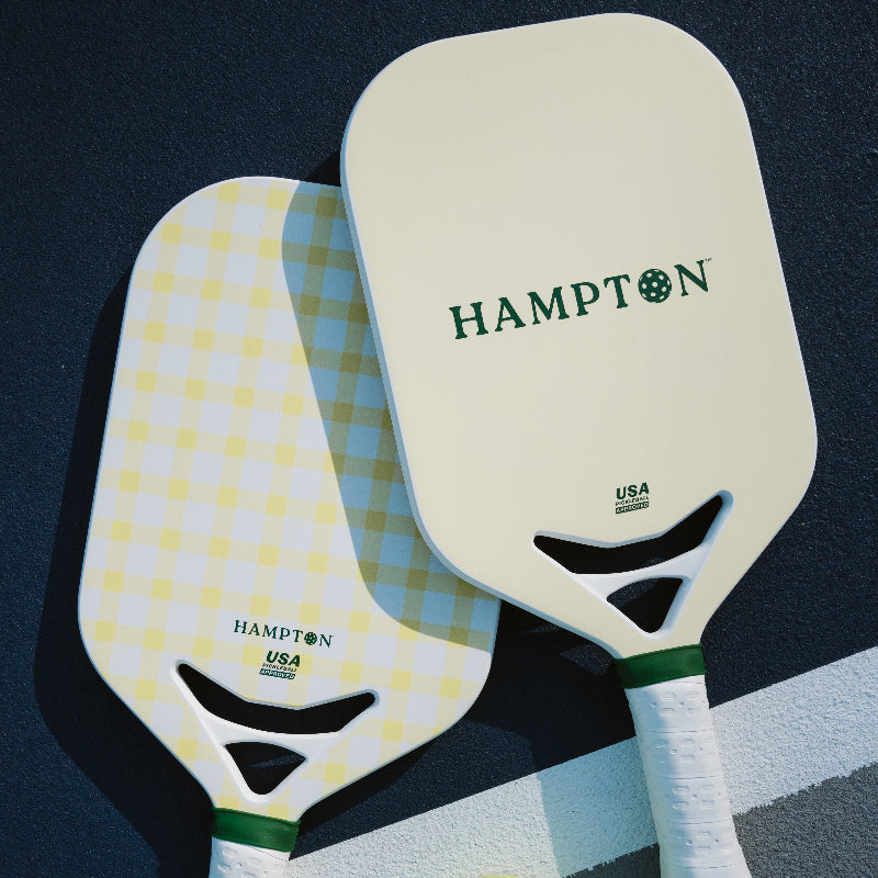 Two pickleball paddle featured laying on a court. One features a cream and yellow plaid pattern. The other is a cream color with a green "HAMPTON" displayed.