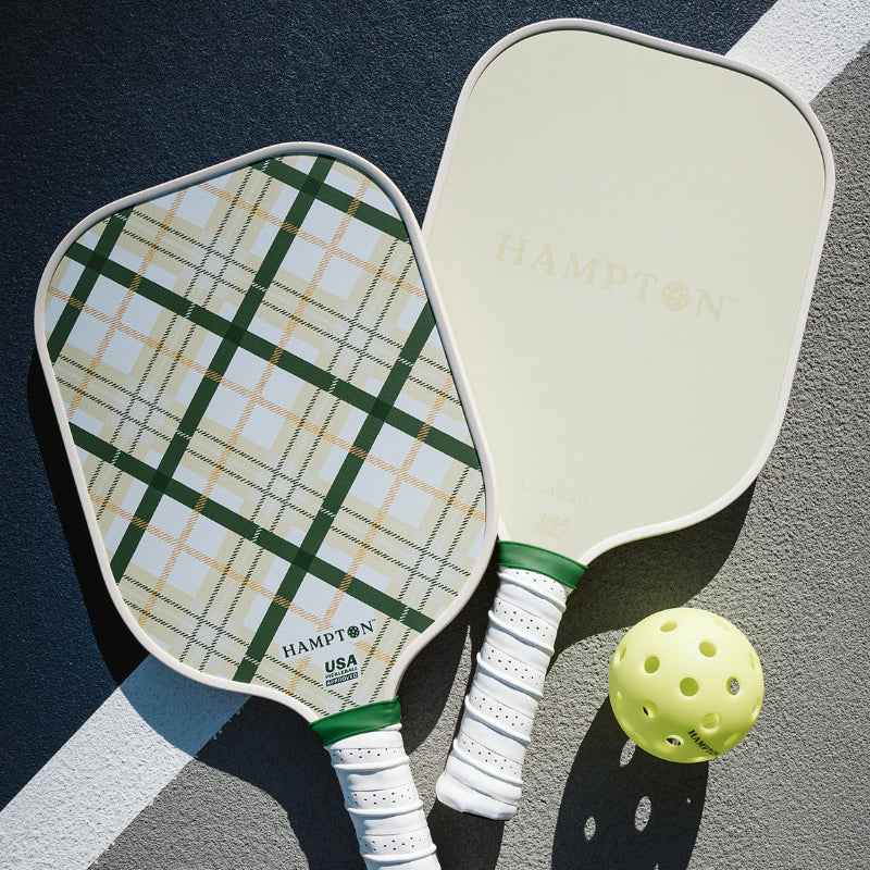 Two pickleball paddles with a green plaid pattern and a yellow pickleball on a court surface. One side is a solid cream color with the word "HAMPTON" on it.