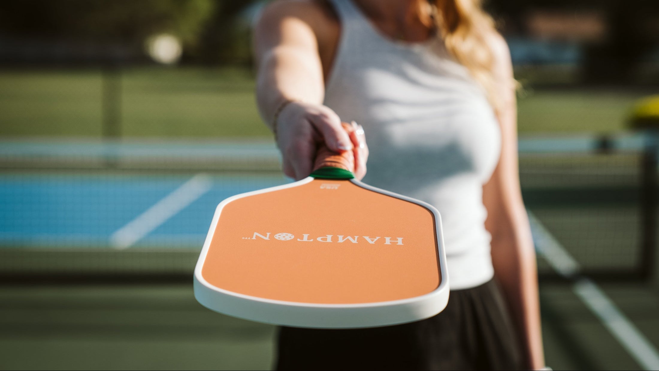 Middle-age woman holding a Hampton Paddles pickleball paddle. She is pointing it at the camera while standing on a pickleball court.