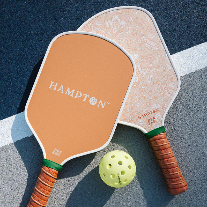 Two orange and white pickleball paddles with green and brown handles, one with the brand name 'HAMPTON' visible, and a yellow pickleball in front of them, placed on a blue and gray surface.